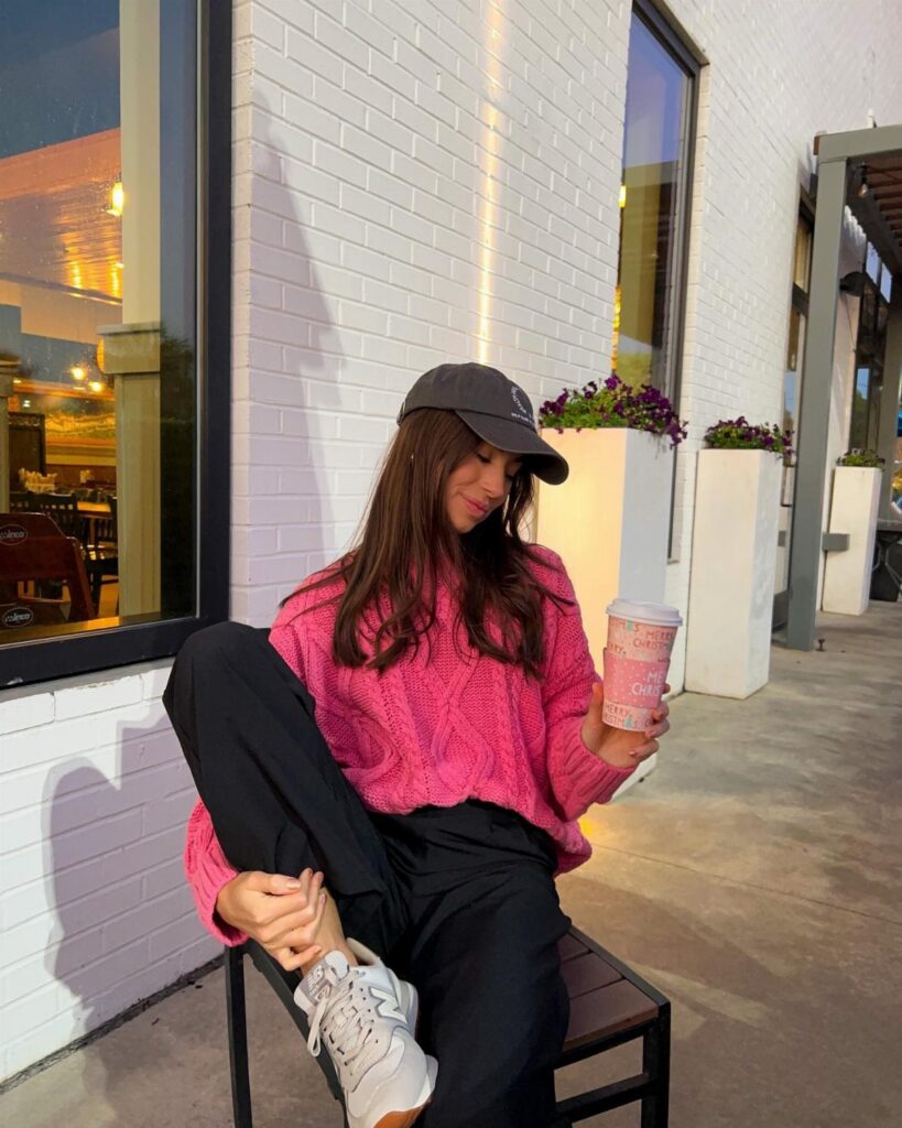 A woman in a pink chunky knit sweater sitting on a bench holding a coffee cup.