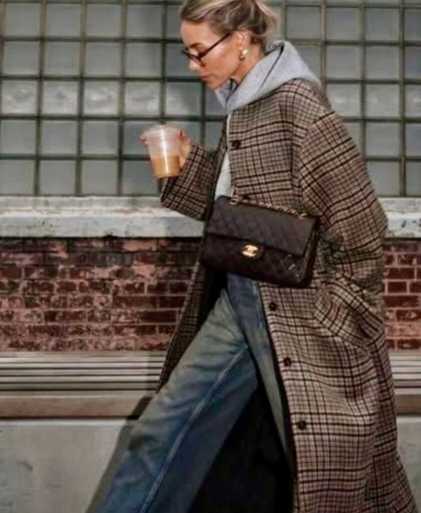 A woman in a plaid coat, grey hoodie, and blue jeans walking with a handbag.