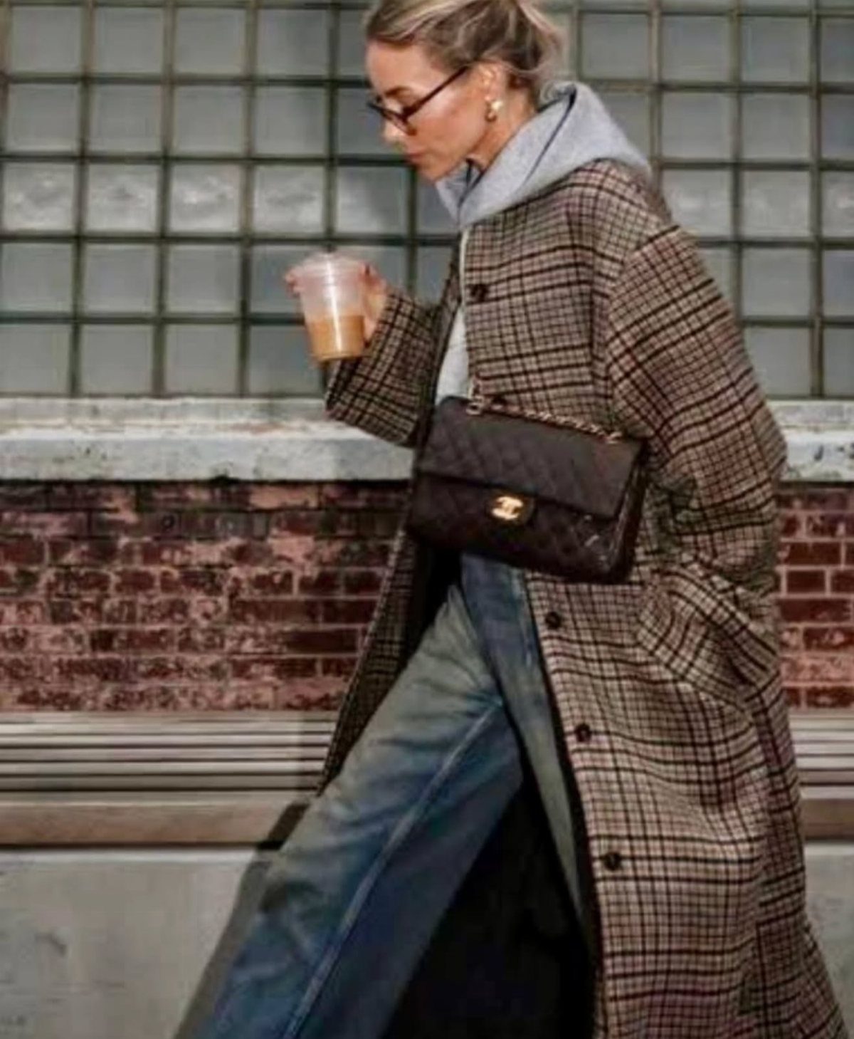 A woman in a plaid coat, grey hoodie, and blue jeans walking with a handbag.