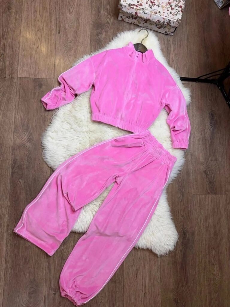 Pink velour tracksuit jacket and matching pants laid on a white faux fur rug.